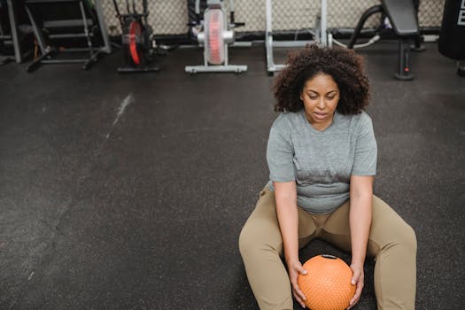 From above of exhausted young obese African American lady with curly hair in activewear sitting on floor with medicine ball in hands while recreating after workout in gym