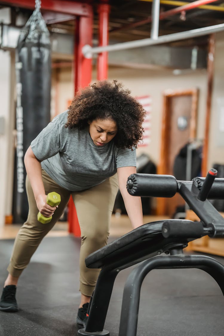 Overweight Black Woman Lifting Dumbbell While Leaning On Gym Equipment