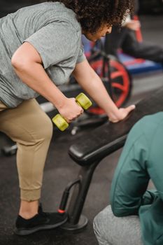 Crop anonymous African American female working out with dumbbell while unrecognizable coach supporting in gym