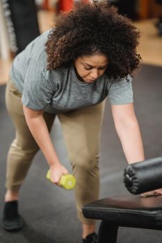 Full body of concentrated African American female lifting dumbbell on blurred background of gym
