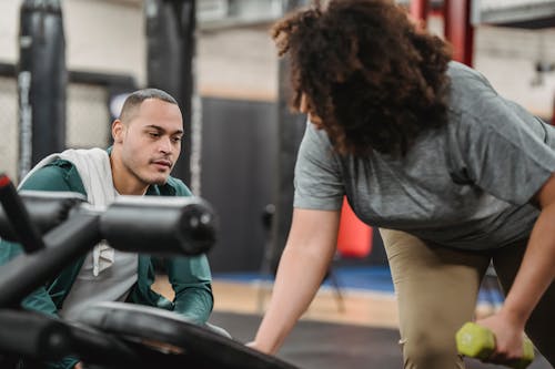 Experienced instructor watching anonymous African American female lifting dumbbell on blurred background of gym