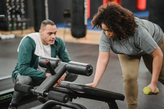 Focused instructor helping plus size African American female doing exercises with dumbbell near gym equipment