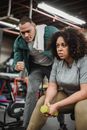 Serious African American female sitting with dumbbell and listening to instruction of personal trainer in gym
