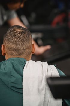 Back view of unrecognizable male with towel on shoulder sitting and resting after training in gym