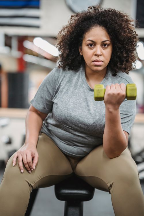 Free Curvy female sitting on bench and doing dumbbell curl during training in gym looking at camera Stock Photo