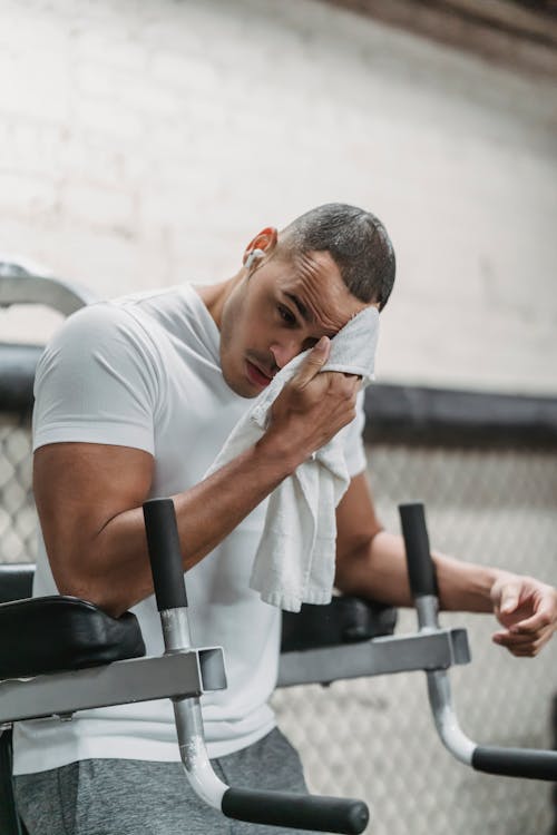 Young male wiping forehead with towel during rest after hard workout in contemporary fitness center