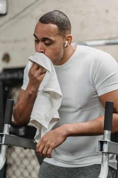 Strong sportsman resting after workout while listening music in earbuds and wiping face with towel in gym