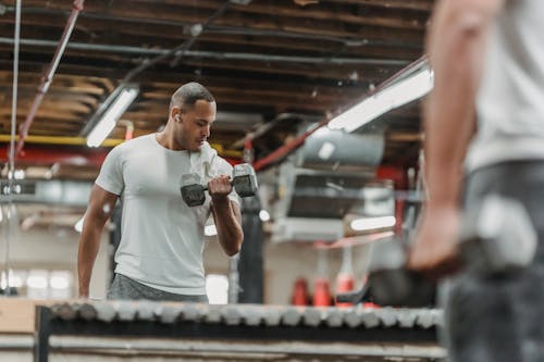 Crop serious male athlete standing in front of mirror and exercising with heavy dumbbells