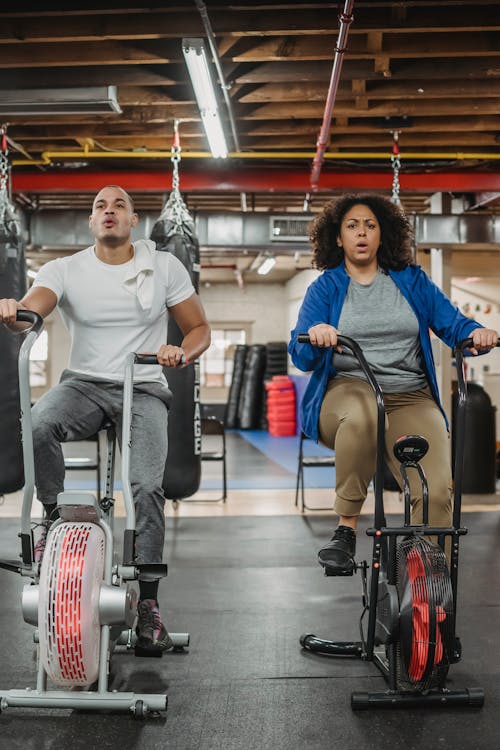 Full body of strong male trainer and plus size female sitting on exercise bikes in spacious modern gym