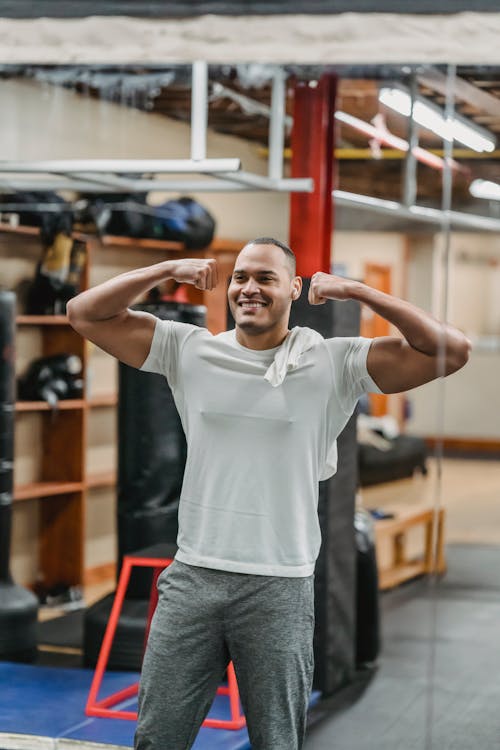 Smiling sportive ethnic male in active wear standing in double biceps pose against mirror in modern gym