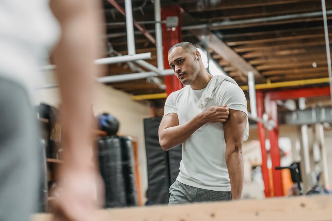 Pensive muscular ethnic sportsman in activewear looking at biceps while reflecting in gym mirror and working out