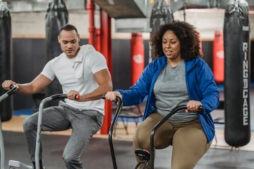 Plus sized African American female in sportswear exercising on stationary bicycle near fit muscular male coach in modern gym