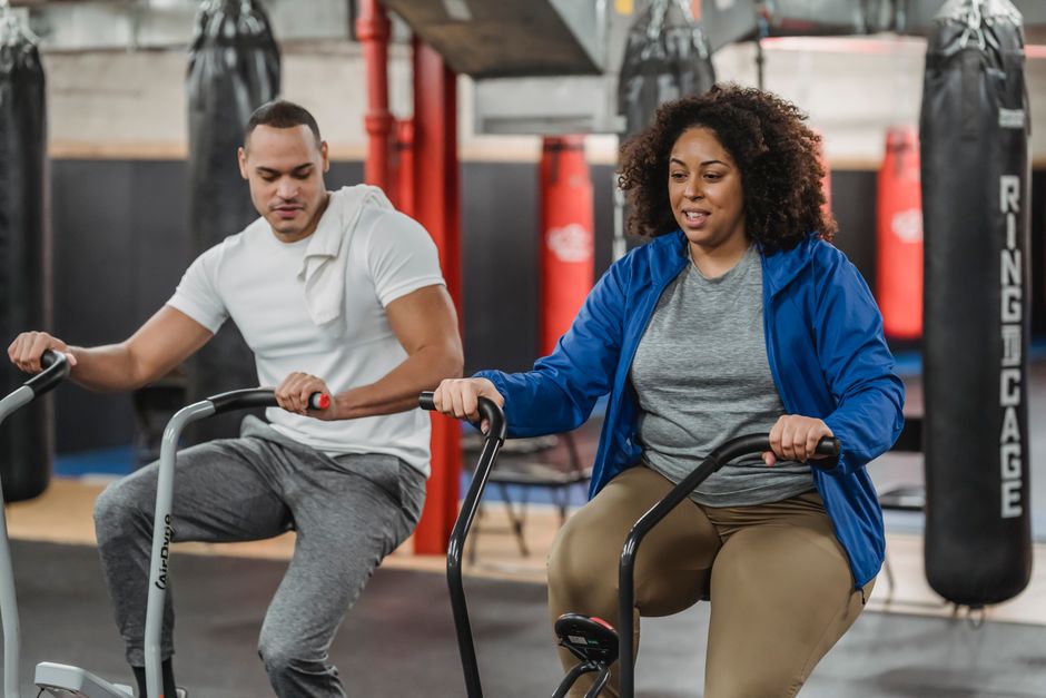 Plus sized African American female in sportswear exercising on stationary bicycle near fit muscular male coach in modern gym