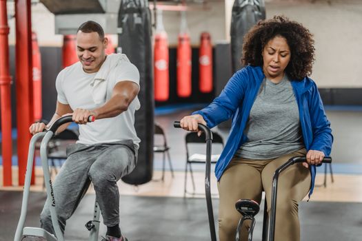 Determined plus sized African American female in sportswear and fit smiling sportsman riding stationary bicycles together in contemporary gym