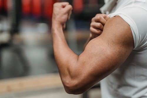 Crop anonymous sportsman in white shirt demonstrating biceps while training in modern gym