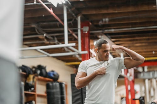 Concentrated young ethnic sportsman in activewear and earbuds looking at biceps while working out in modern gym