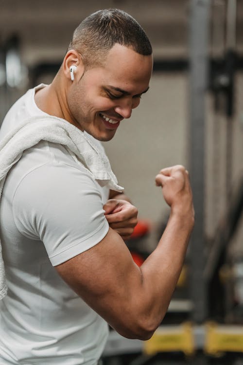 Side view happy ethnic sportsman in activewear and earbuds showing strong bicep while working out in contemporary gym