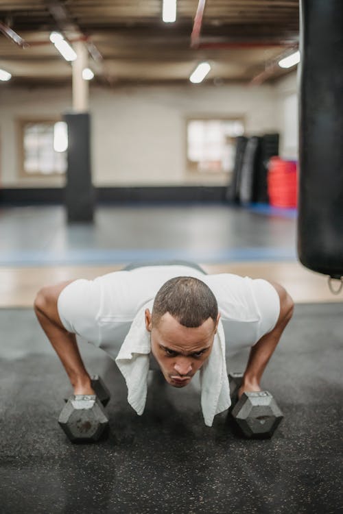 Strong ethnic sportsman in activewear doing pushups with dumbbells in modern equipped fitness club