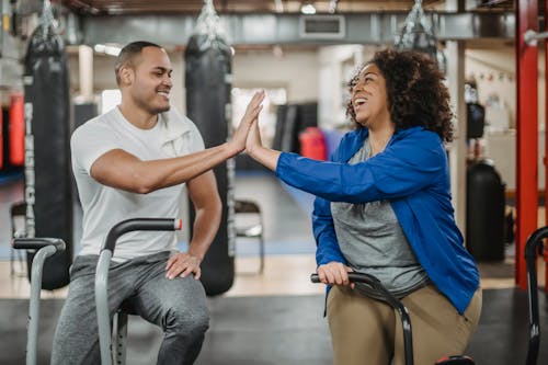 Happy plump African American female trainee and muscular smiling coach clapping each others hands while sitting on stationary bicycles in modern gym