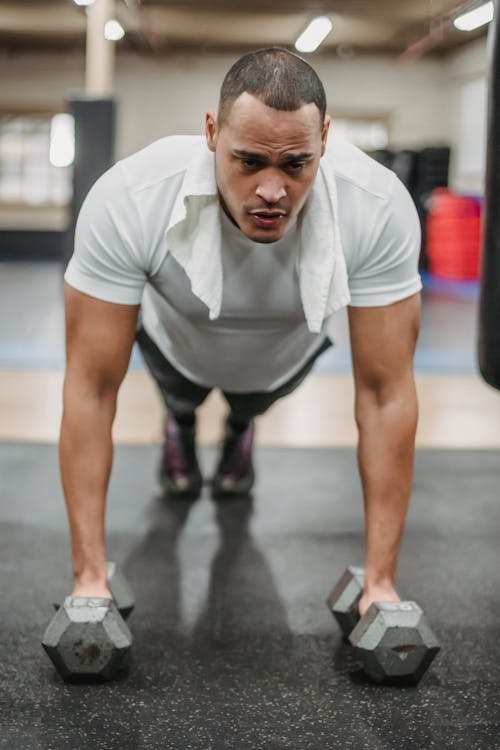Full body powerful concentrated ethnic sportsman in activewear performing pushing ups with heavy dumbbells during intense workout in contemporary fitness club