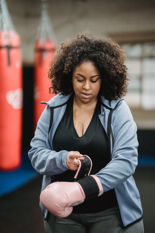 Free Positive plus sized African American female putting on pink boxing gloves before training in modern gym Stock Photo