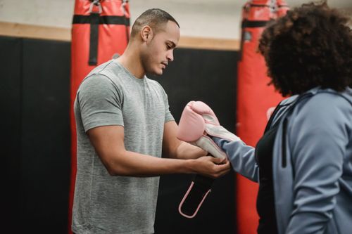 Free Serious ethnic male instructor standing near punching bags and putting on boxing gloves on overweight female before workout Stock Photo