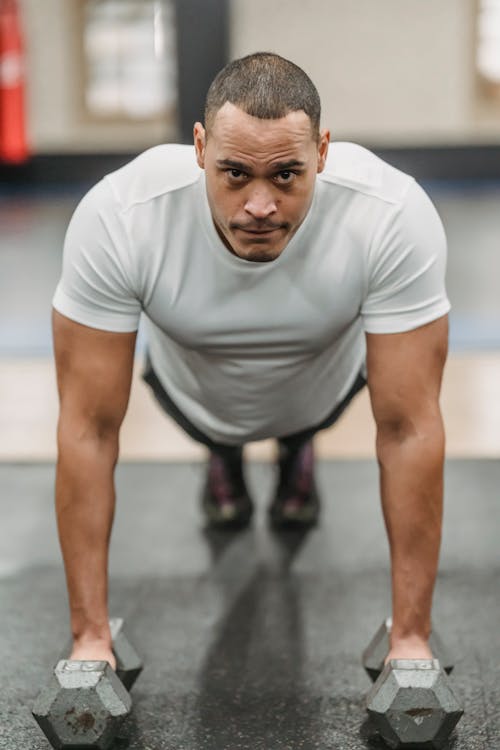 Full body of focused ethnic male standing in plank position with dumbbells while exercising in gym