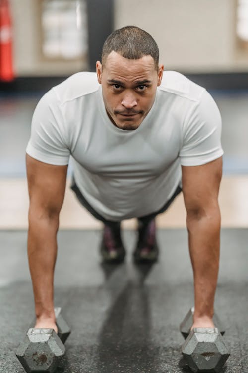 Free Full body of focused ethnic male standing in plank position with dumbbells while exercising in gym Stock Photo