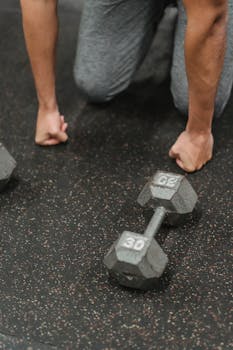 A man kneels on the gym floor beside a pair of 30-lb dumbbells, taking a break from exercise.