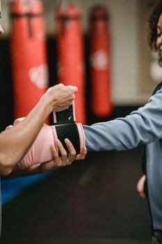 Side view of crop personal trainer preparing African American female for boxing training in gym with punching bags