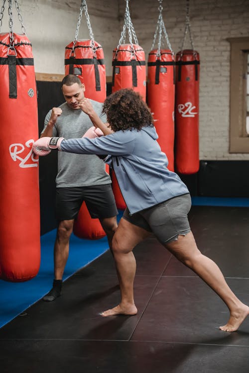 Full body of strong plus size African American female in boxing gloves hitting punching bag during training with personal instructor showing technique