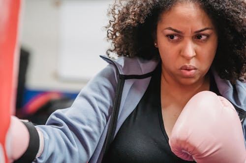 Crop plump African American woman in boxing gloves doing hit of punching bag during hard training in gym