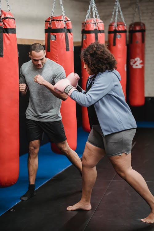 Strong ethnic man showing stance to overweight African American woman in boxing gloves hitting punching bag