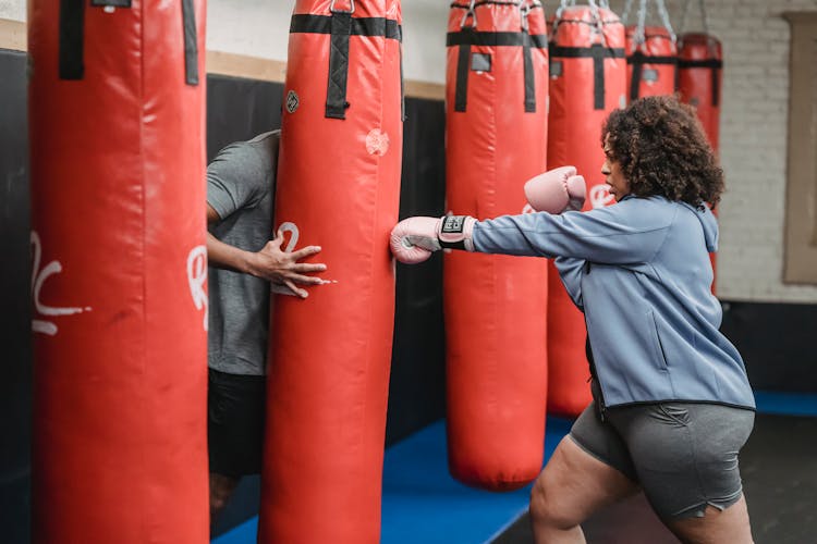 Black Woman Hitting Punching Bag During Workout With Trainer