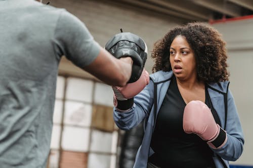 Active African American female in boxing gloves practicing punches while training with unrecognizable personal coach during boxing workout in gym