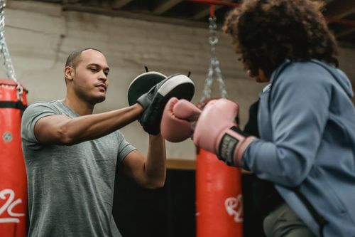 Free Strong African American personal instructor with boxing paws training anonymous female with gloves while practicing in gym with punching bag Stock Photo