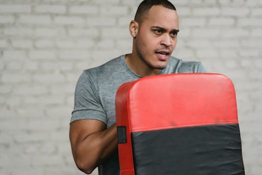 Crop muscular ethnic sportsman with focus mitt looking away in gymnasium on blurred gray background