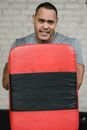 Adult ethnic male athlete behind focus mitt looking at camera in gym on blurred background