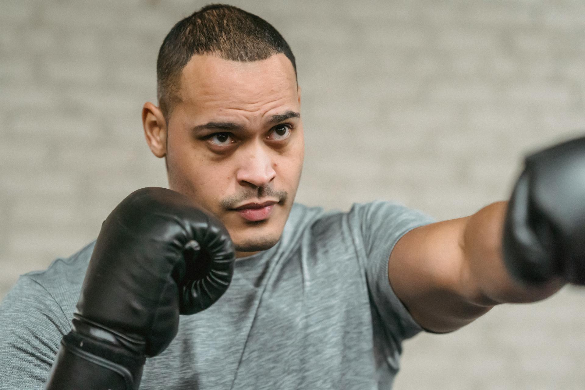 Strong ethnic sportsman in boxing gloves performing punching technique while looking forward during training on blurred gray background