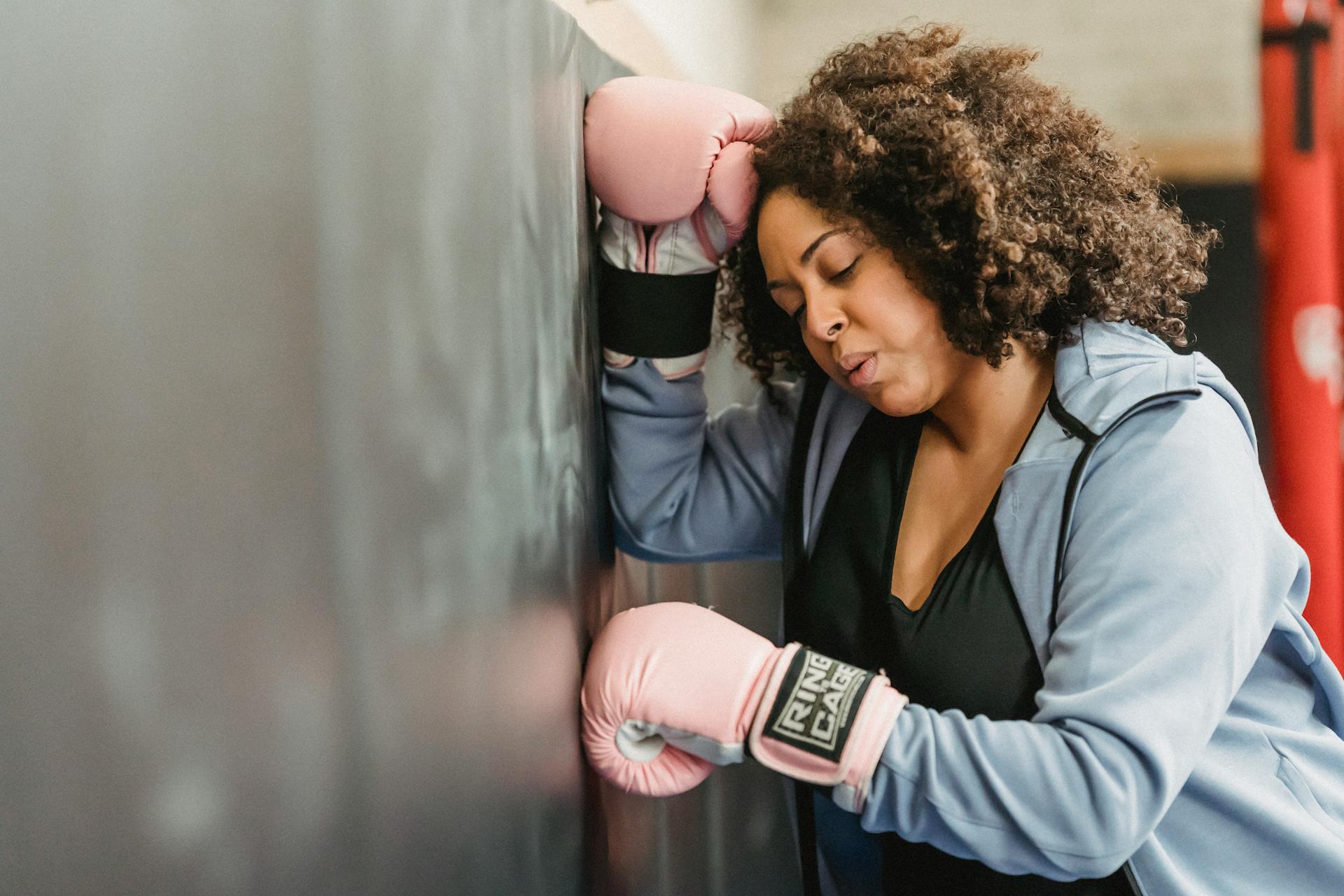 Sidovision av trött ung afroamerikansk kvinnlig kämpe med mörkt lockigt hår i sportkläder och skyddshandskar som lutar sig mot väggen efter boxningsträning i gymmet