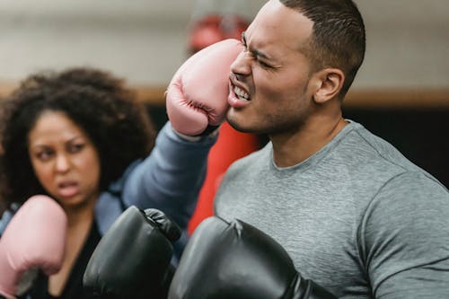 Free Side view of young muscular ethnic male trainer in sportswear and boxing gloves receiving heavy punch on face from serious young African American female during workout Stock Photo