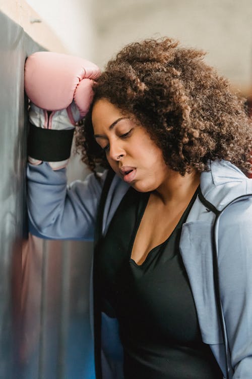 Side view of exhausted young ethnic female fighter with Afro hair in sportswear and boxing gloves leaning on wall and breathing after hard workout