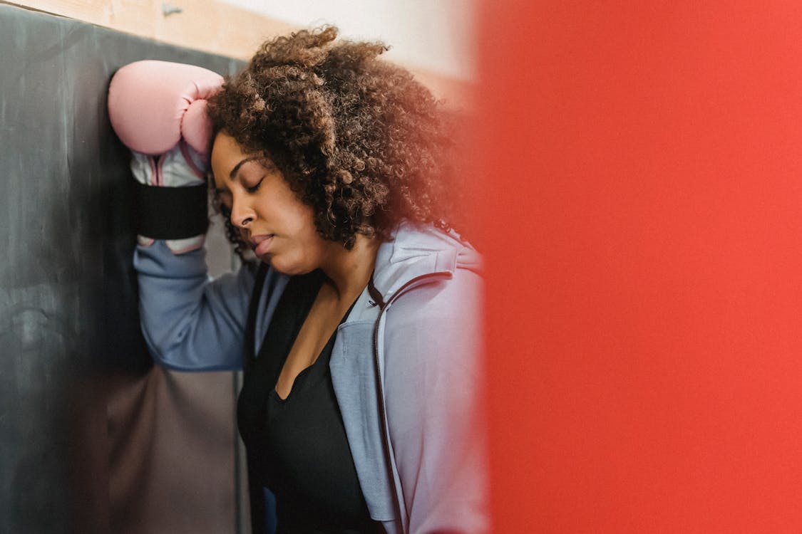 Free Side view of tired young black female with dark curly hair in sportswear and protective gloves leaning on wall with closed eyes after hard training in gym Stock Photo