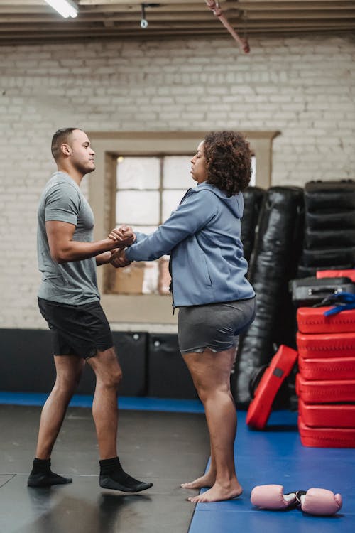 Side view of young concentrated ethnic male trainer stretching hands of tired plus size African American female after boxing workout in gym