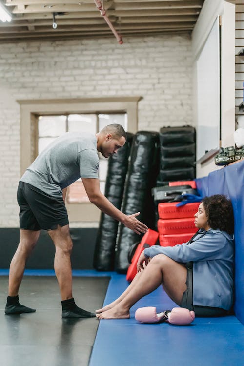 Full body side view of muscular black personal coach reaching out hand to African American female while training in gym