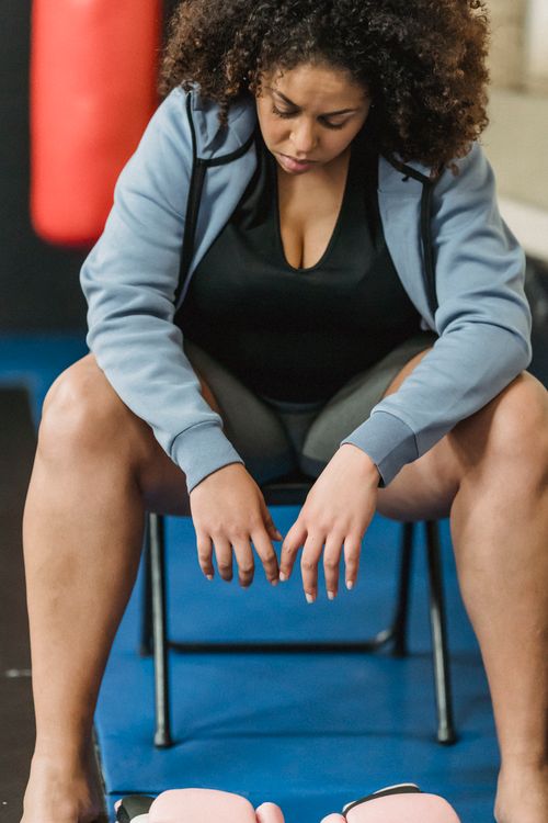 Free Serious overweight African American female in sportswear looking down while sitting on chair on mar with boxing gloves during workout in gym on blurred background Stock Photo