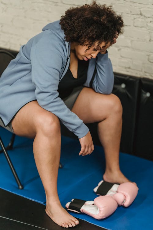 Full body of tired African American female in activewear sitting on chair near boxing gloves during workout in modern gym