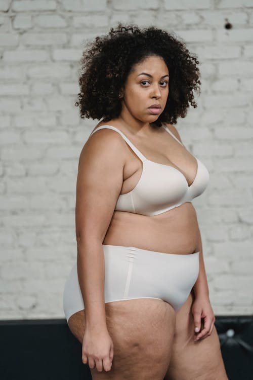 Confident overweight African American woman in bra and panties looking at camera while standing near white brick wall in studio