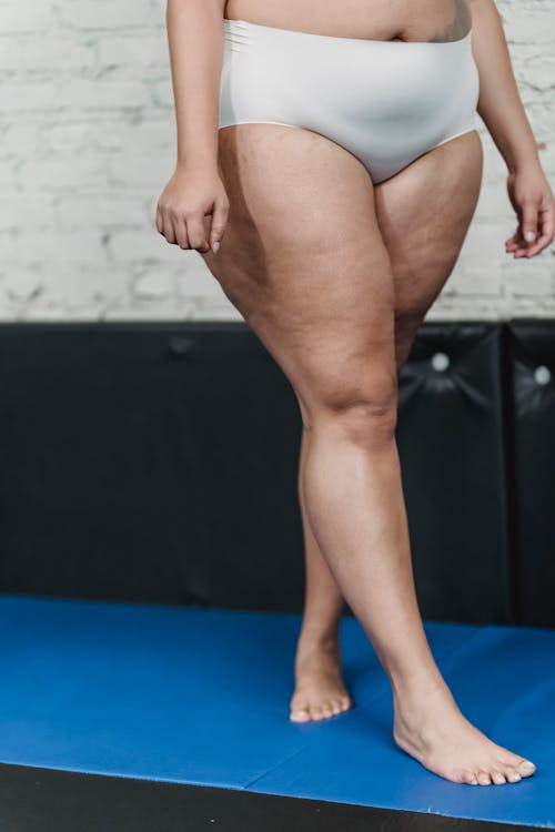 Unrecognizable plus size barefoot female with stretched marks in panties walking on blue mat near white brick wall in gym