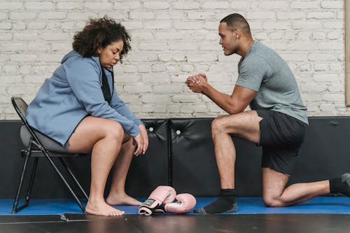 Free Side view of muscular African American male trainer explaining technique of exercise to plump black woman while sitting on mat with boxing gloves Stock Photo